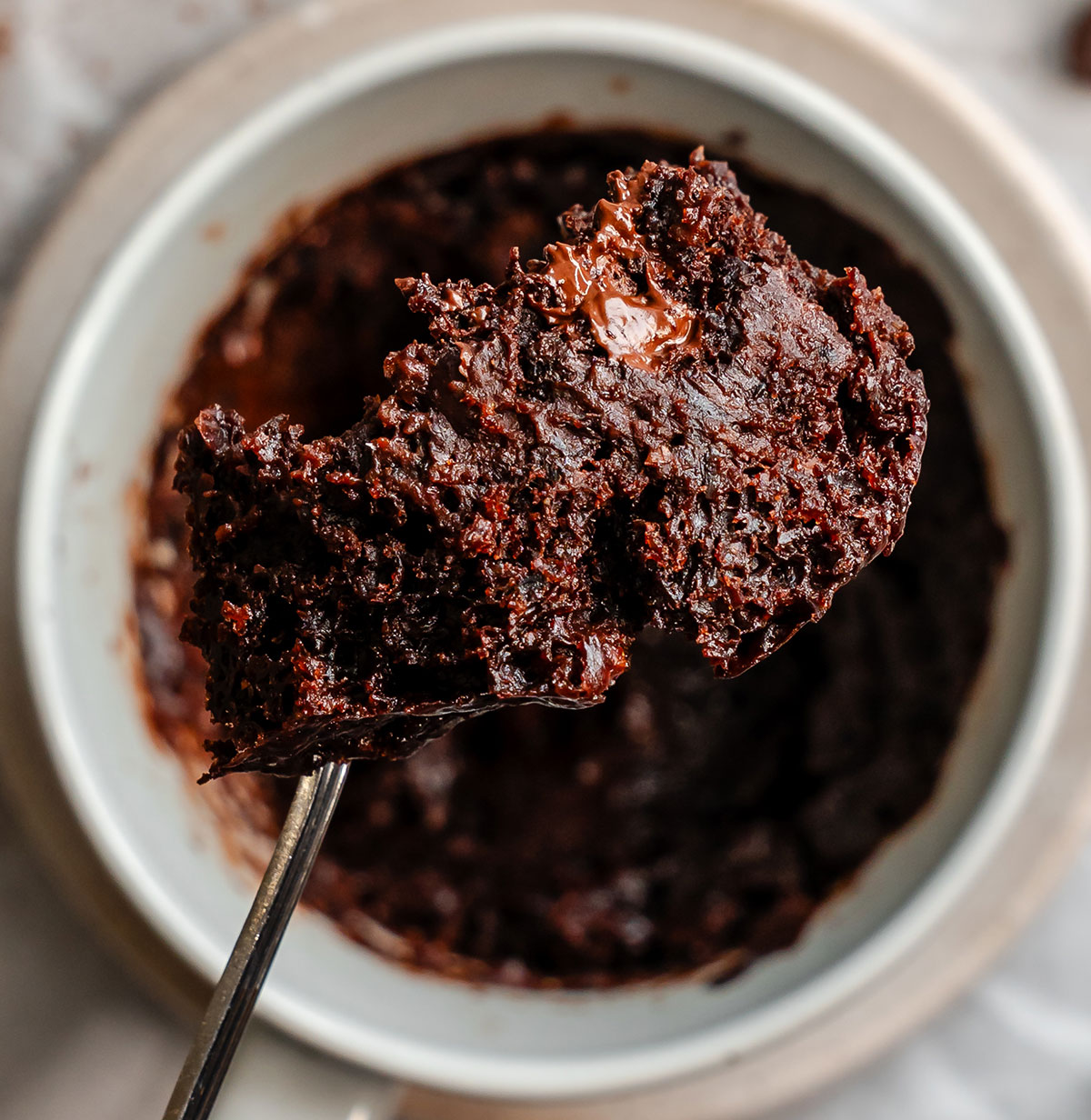 A close-up of a spoon holding a portion of chocolate mug cake above a white ramekin filled with the same dessert.