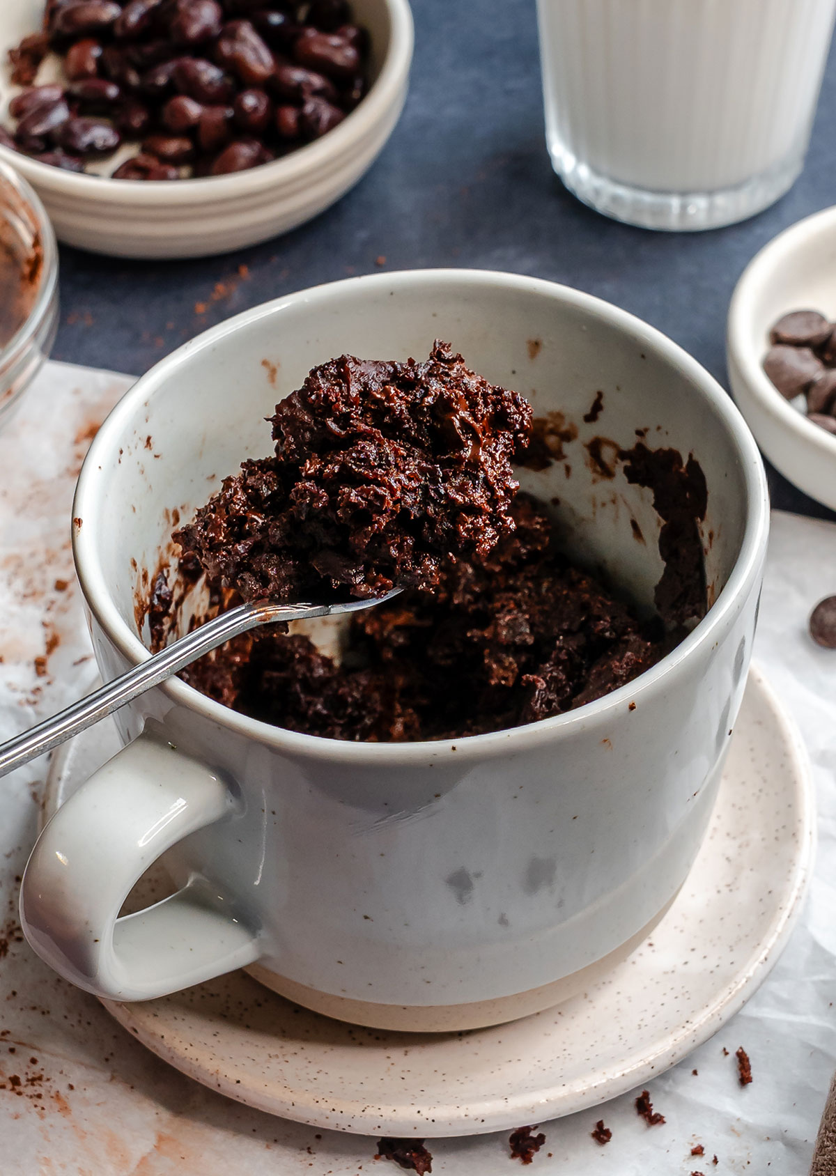 A spoonful of chocolate mug cake is held above a gray mug on a saucer, with bowls of chocolate and a glass of milk in the background.