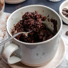 A spoon holds a portion of chocolate mug cake above a mug, with chocolate chips and cocoa powder in small bowls nearby.