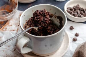 A spoon holds a portion of chocolate mug cake above a mug, with chocolate chips and cocoa powder in small bowls nearby.