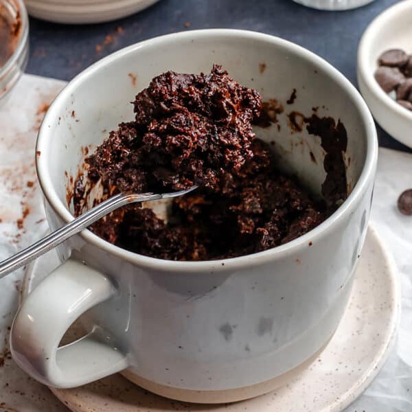 A spoon holds a portion of chocolate mug cake above a mug, with chocolate chips and cocoa powder in small bowls nearby.