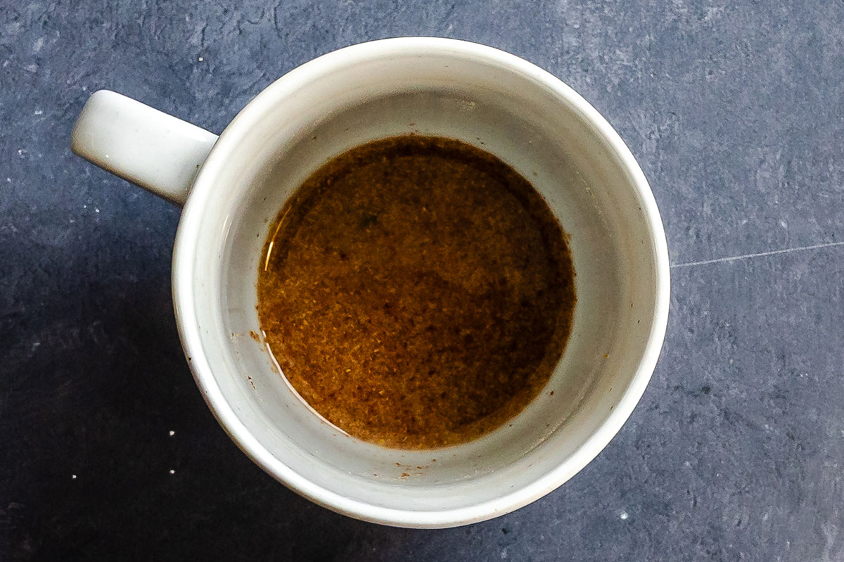 A white mug filled with a small amount of brown liquid sits on a dark, textured surface.