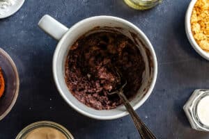 A white mug containing a dark, mixed batter with a spoon inside, surrounded by small bowls of ingredients on a dark countertop.