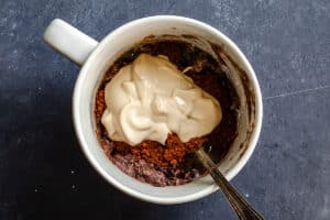 A white mug with a spoon contains cocoa powder, a creamy topping, and a mixture of ingredients, placed on a dark surface.
