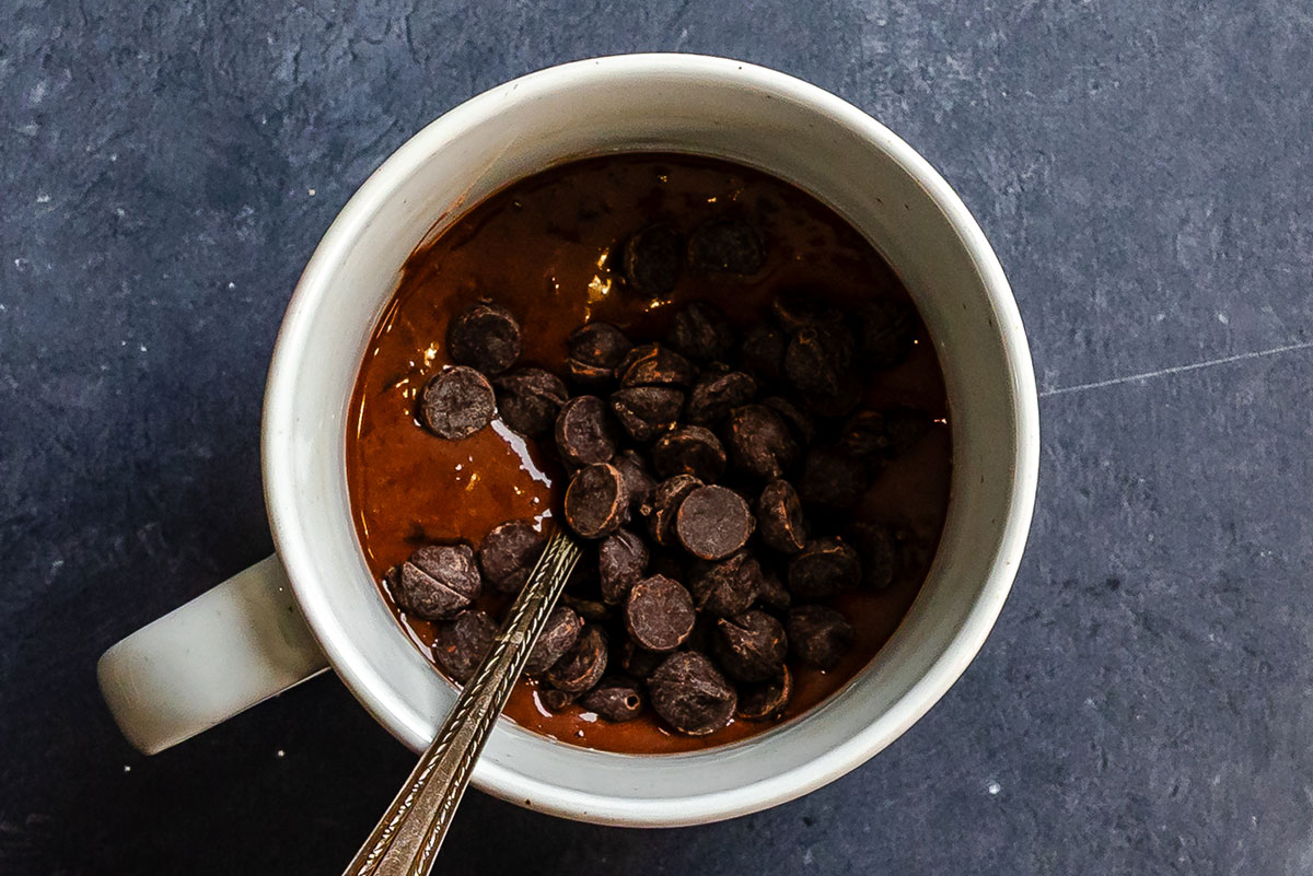 A ceramic mug filled with chocolate batter and topped with chocolate chips, with a spoon inside, on a dark surface.