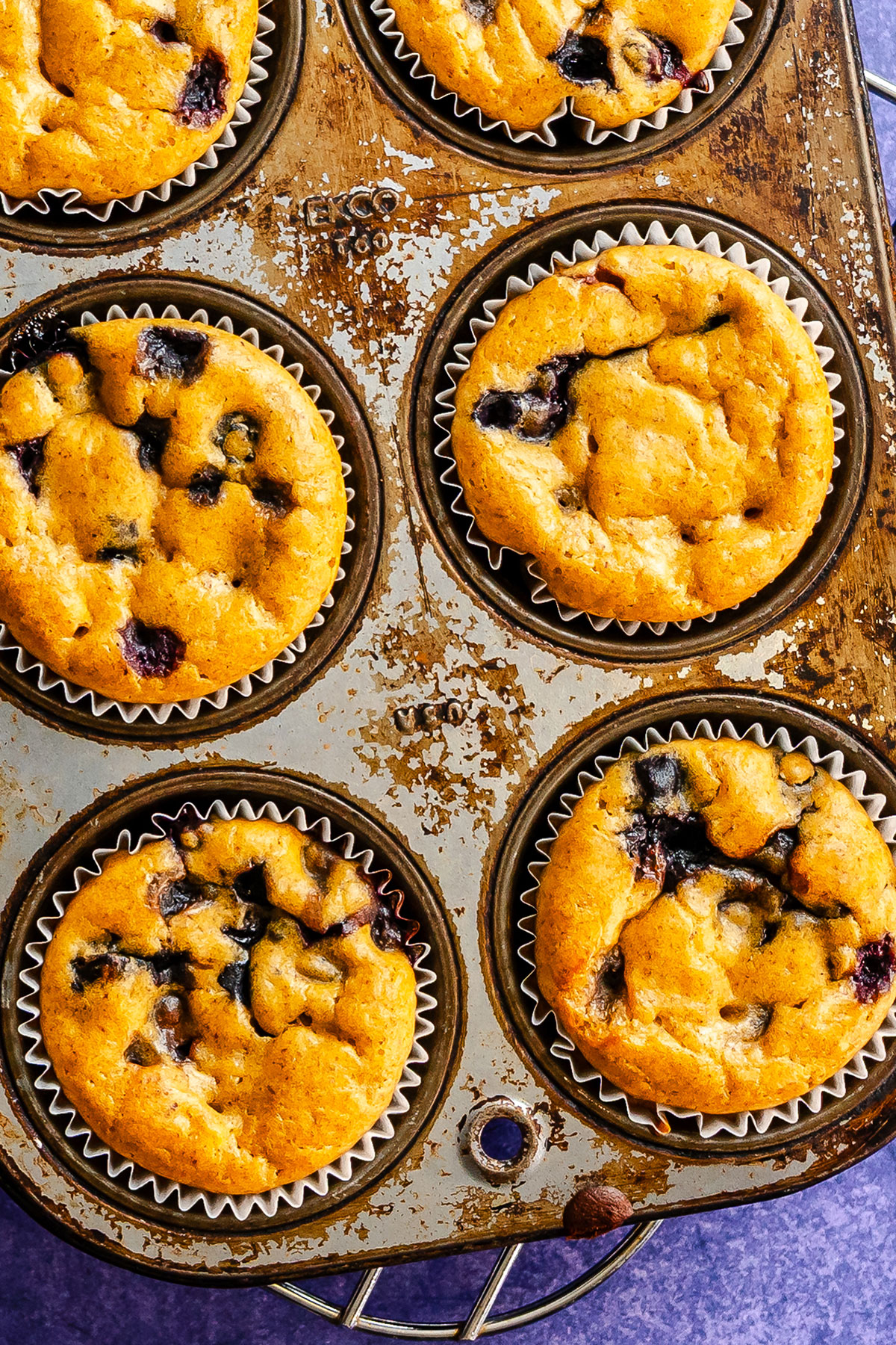 A muffin tin holds six baked blueberry chickpea muffins in paper liners. The tin appears slightly worn and sits on a purple surface.