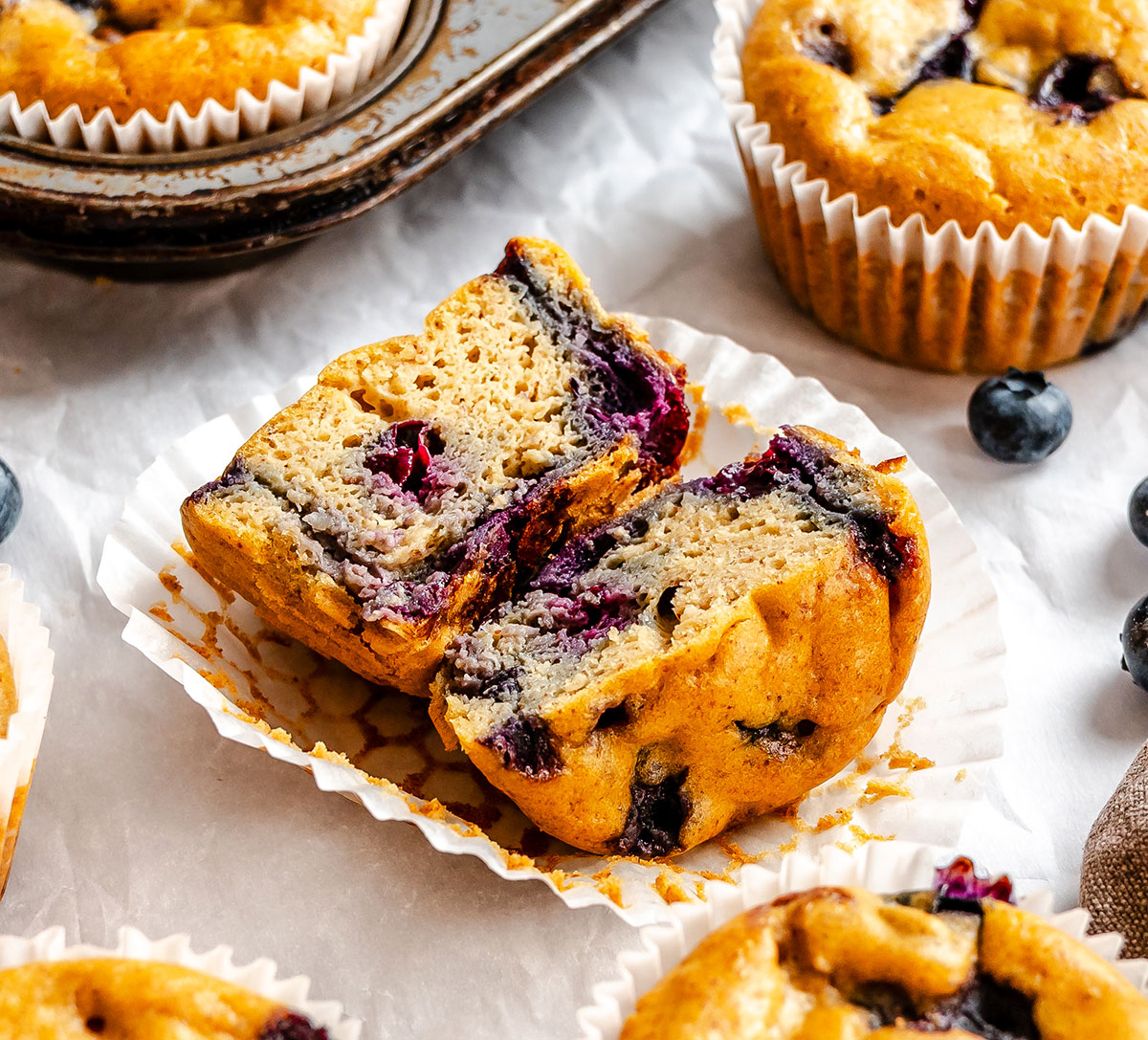 A blueberry muffin cut in half sits on a white muffin liner, surrounded by whole blueberry muffins and a metal muffin tin.