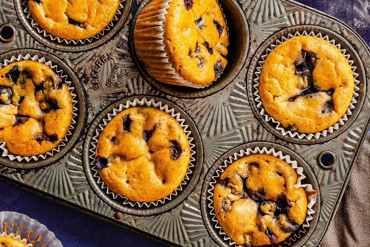 A muffin tin filled with six baked blueberry muffins, five in the tin and one partially lifted out, all in white paper liners.