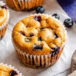 Close-up of a chickpea blueberry muffin in a white paper liner, with visible blueberries, on parchment paper with loose blueberries nearby.