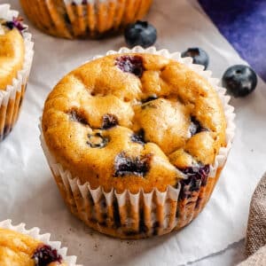 Close-up of a chickpea blueberry muffin in a white paper liner, with visible blueberries, on parchment paper with loose blueberries nearby.