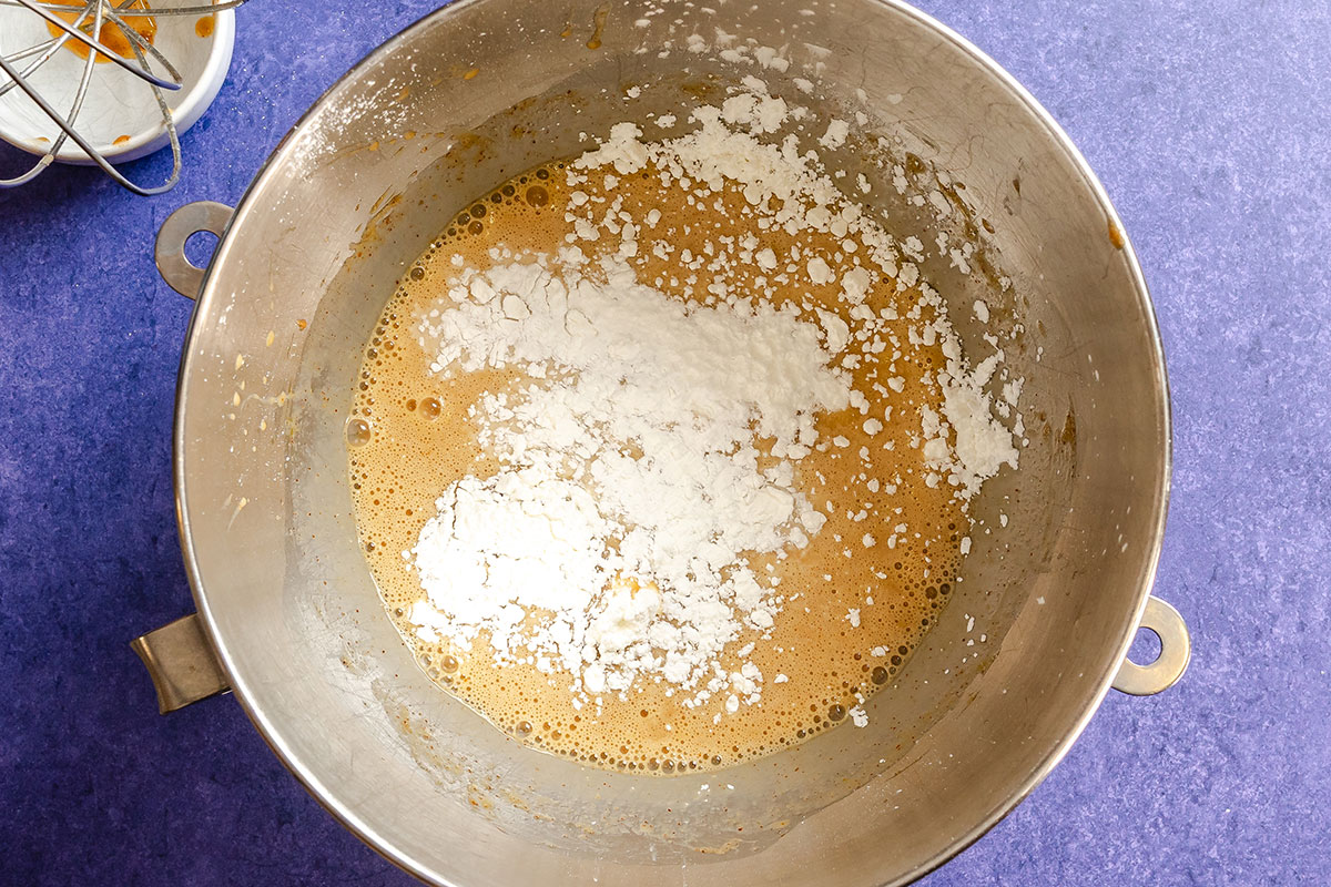 A metal mixing bowl contains a brown batter with a layer of white flour on top, set on a blue countertop.