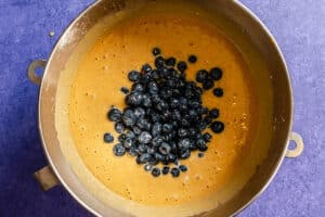Batter with fresh blueberries sits in a large metal mixing bowl on a purple countertop, ready to be mixed.