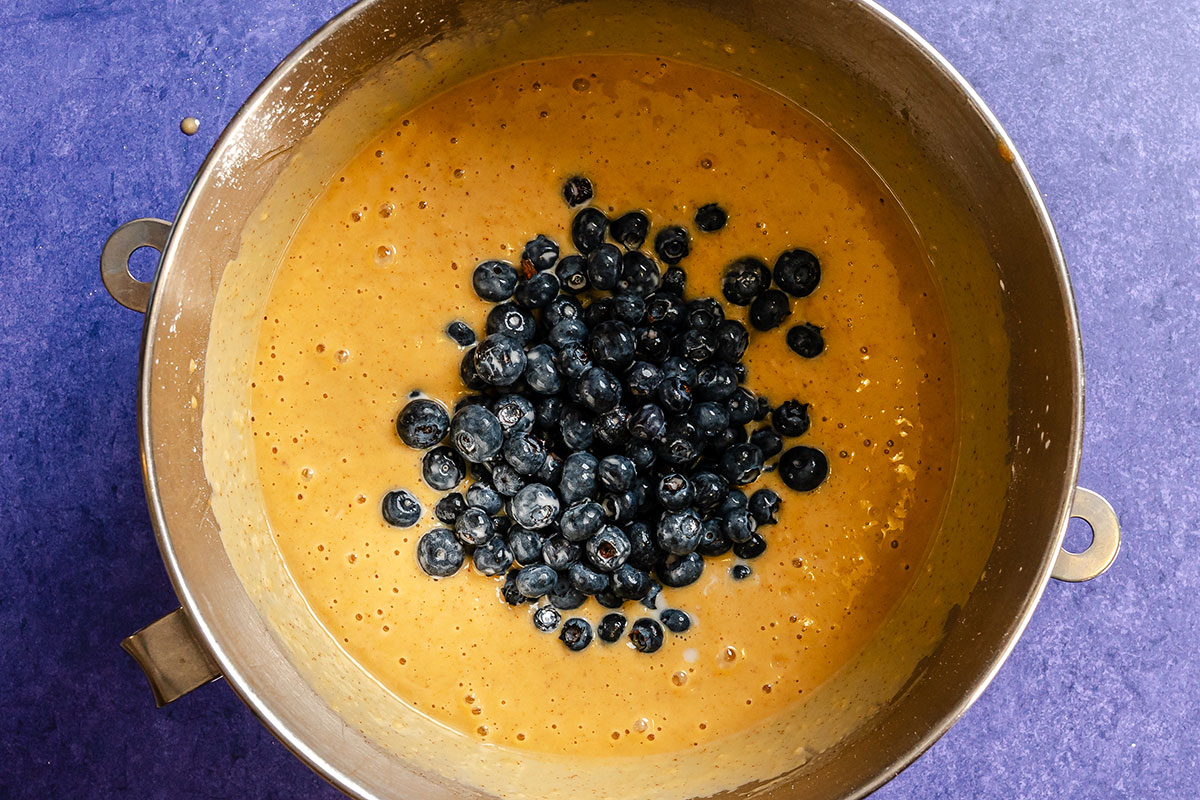 Batter with fresh blueberries sits in a large metal mixing bowl on a purple countertop, ready to be mixed.