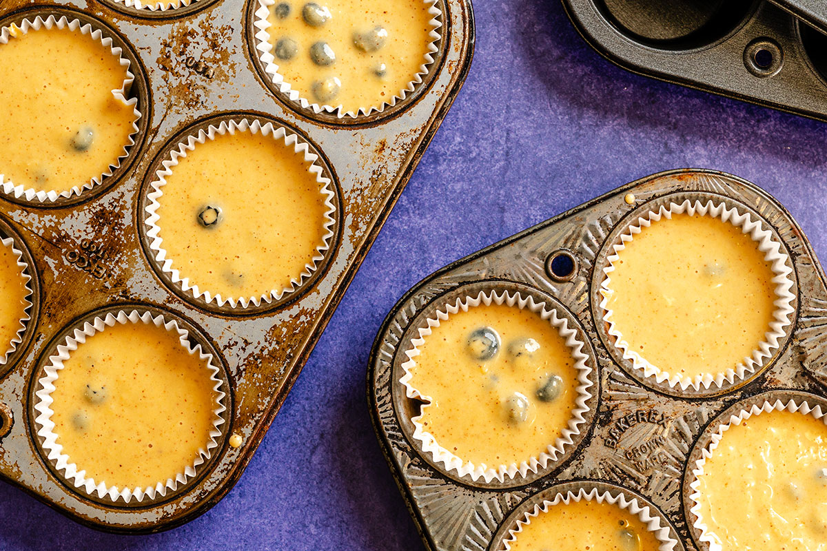 Three muffin trays filled with yellow batter, some with blueberries, are arranged on a purple surface.