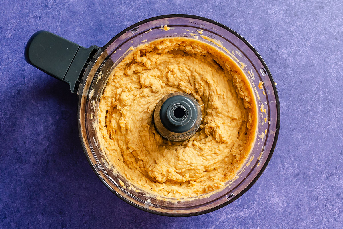 A food processor filled with blended hummus sits on a purple countertop, viewed from above.