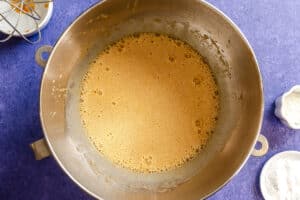 A large metal mixing bowl containing a foamy light brown liquid batter sits on a blue countertop, with small bowls of flour and other ingredients nearby.