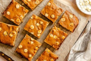 Nine rectangular blondies with white chocolate chips are arranged on parchment paper, viewed from above. A plate of loose white chocolate chips and a striped cloth are partially visible nearby.