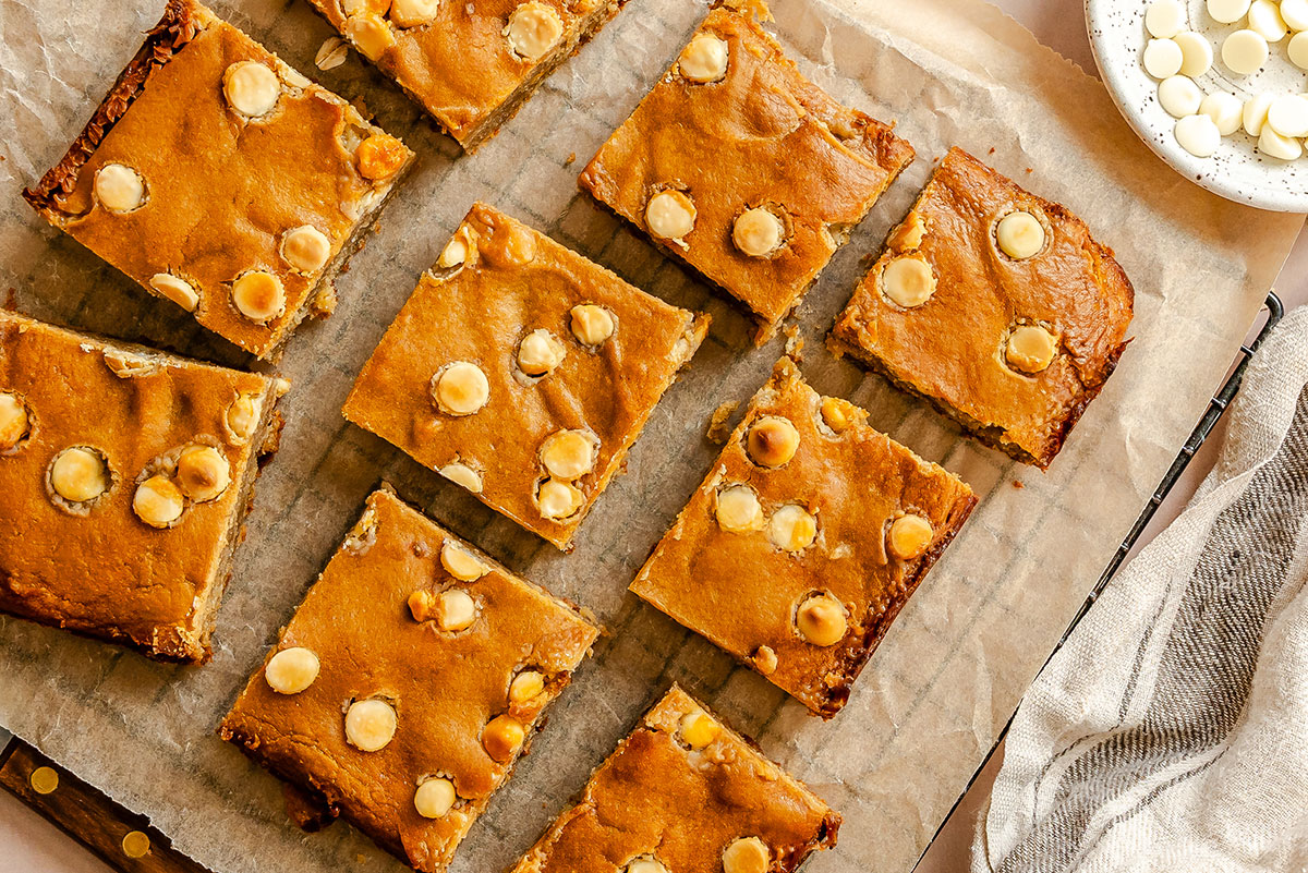 Nine rectangular blondies with white chocolate chips are arranged on parchment paper, viewed from above. A plate of loose white chocolate chips and a striped cloth are partially visible nearby.