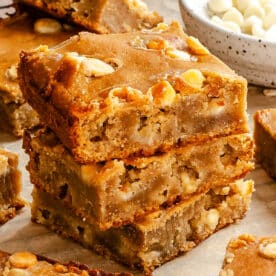 Three stacked butter bean blondies with white chocolate chips and chopped nuts on parchment paper, with a bowl of white chocolate chips in the background.