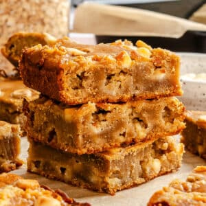 Three blondie dessert bars stacked on top of each other, showing a moist, dense texture with visible white chocolate chunks and nuts, set on parchment paper with more bars in the background.