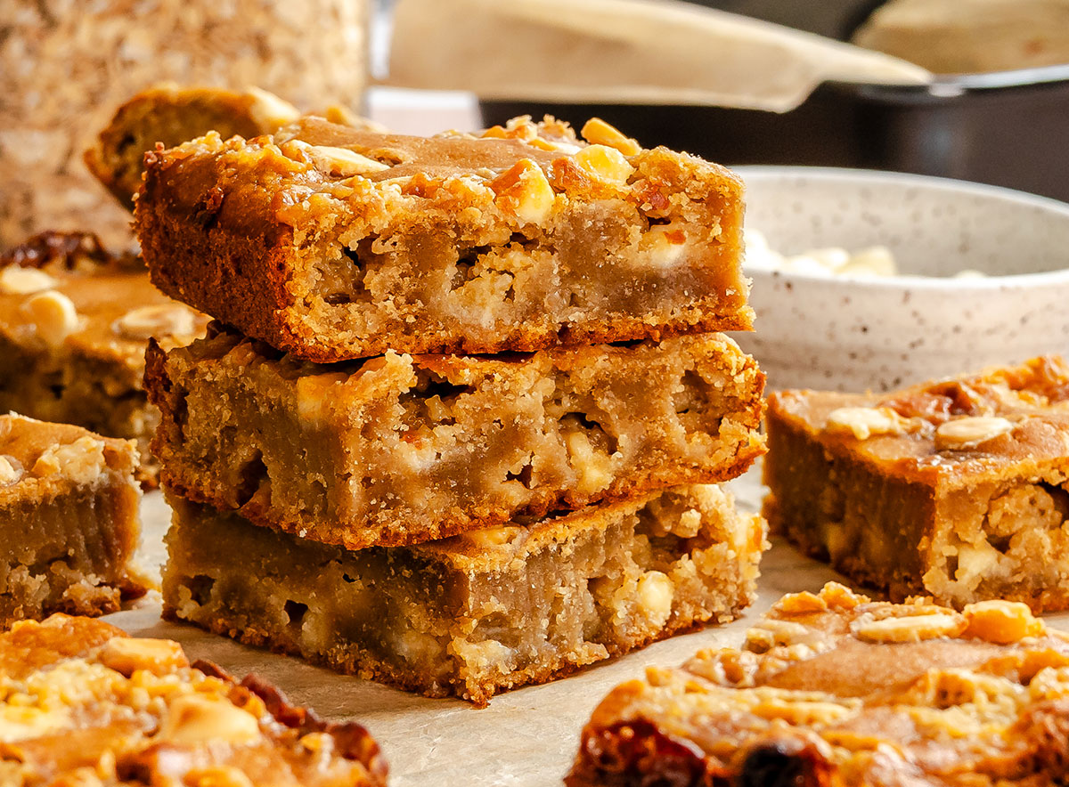 Three bean blondie bars with white chocolate chips and nuts are stacked on a piece of parchment paper, with more blondies and a bowl in the background.