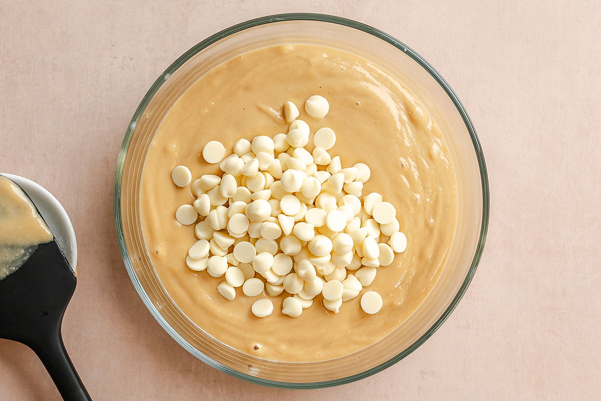 A glass bowl filled with light brown batter, topped with white chocolate chips, sits on a beige surface next to a black spatula coated with batter.