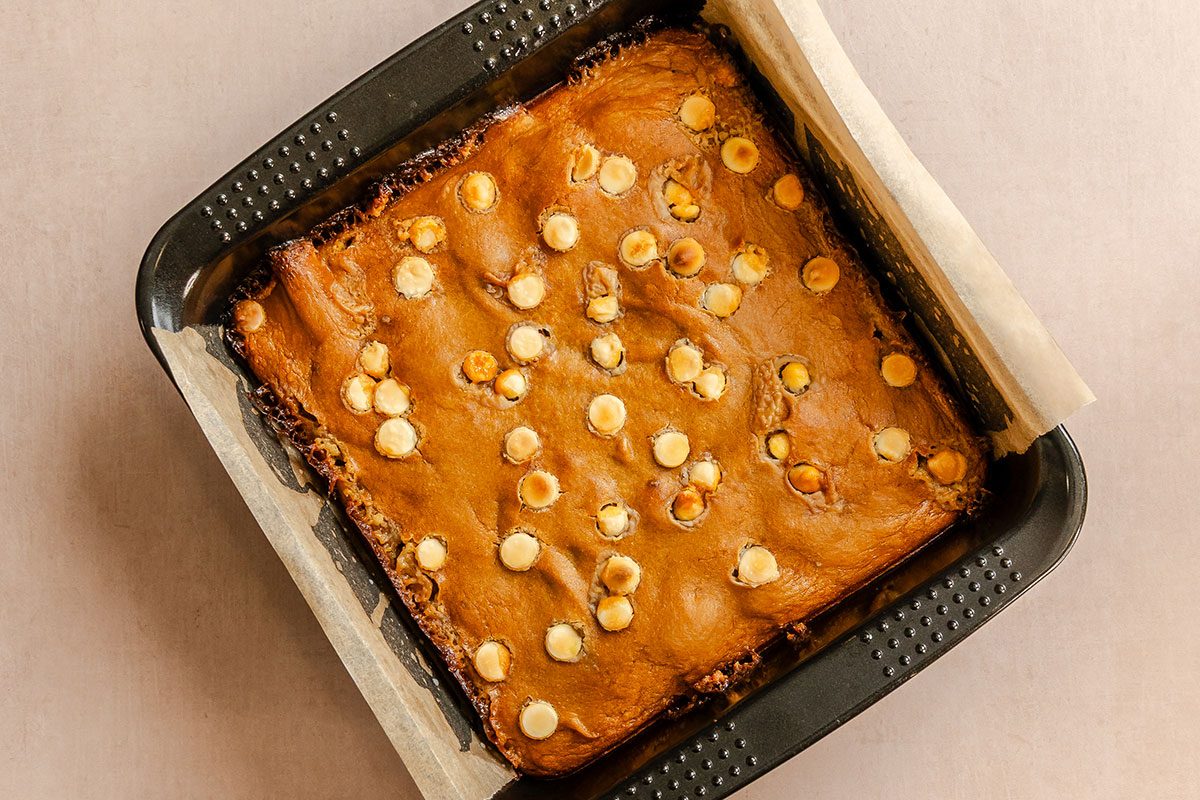 A square pan lined with parchment paper holds baked blondies topped with scattered white chocolate chips.