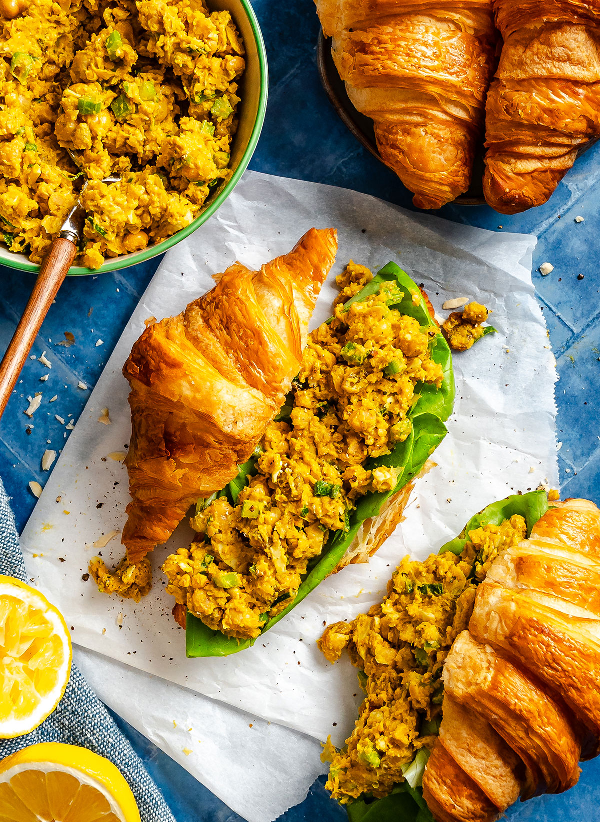 Two croissant sandwiches filled with a yellow chickpea salad and lettuce on parchment paper, with extra chickpea salad and croissants nearby. Lemon halves are also visible.