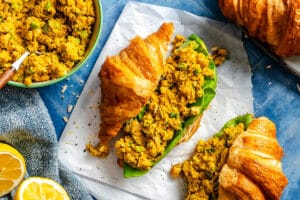 A croissant sandwich filled with curried chickpea salad and lettuce sits on parchment; extra salad, croissants, and lemon halves surround it.