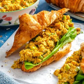 A croissant sandwich filled with leafy greens and a yellow chickpea salad sits on parchment paper, with more chickpea salad and croissants visible in the background.
