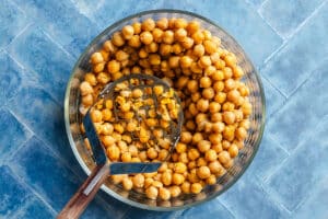 A glass bowl of chickpeas with a metal potato masher, some chickpeas partially mashed, on a blue tiled surface.