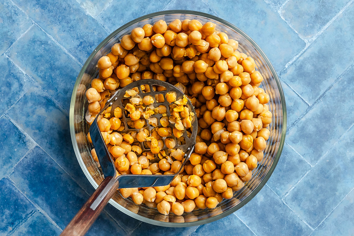 A glass bowl of chickpeas with a metal potato masher, some chickpeas partially mashed, on a blue tiled surface.
