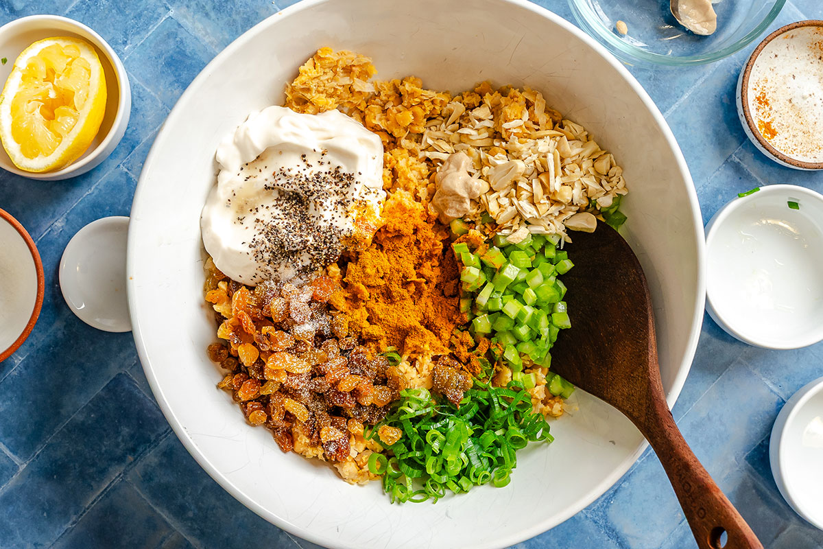 A white bowl contains chopped celery, oats, raisins, green onions, chickpeas, mayonnaise, turmeric, and chia seeds, with a wooden spoon on the side. Surrounding bowls hold lemon and condiments.
