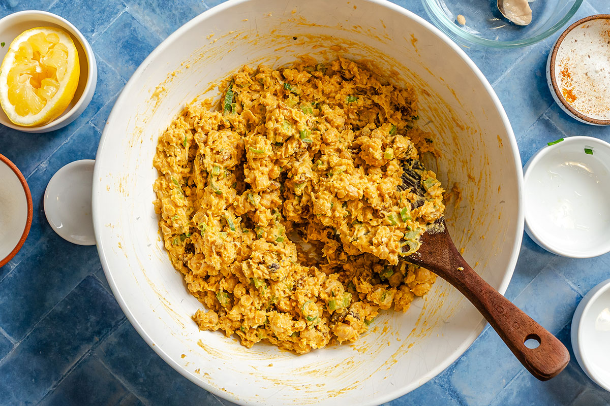 Large white bowl filled with a mashed chickpea mixture being stirred with a wooden spoon; various small bowls with lemon and seasonings surround it on a blue tiled surface.