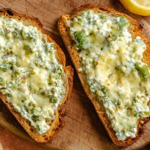 Two slices of toasted bread topped with melted cheese, ricotta, and green olives, served on a wooden board with a lemon wedge and green olives nearby.