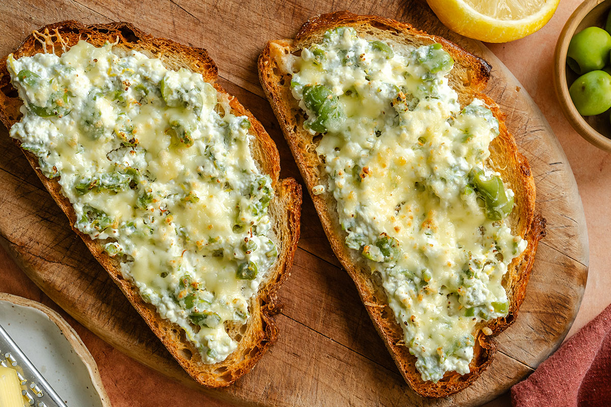 Two slices of toasted bread topped with melted cheese, ricotta, and green olives, served on a wooden board with a lemon wedge and green olives nearby.