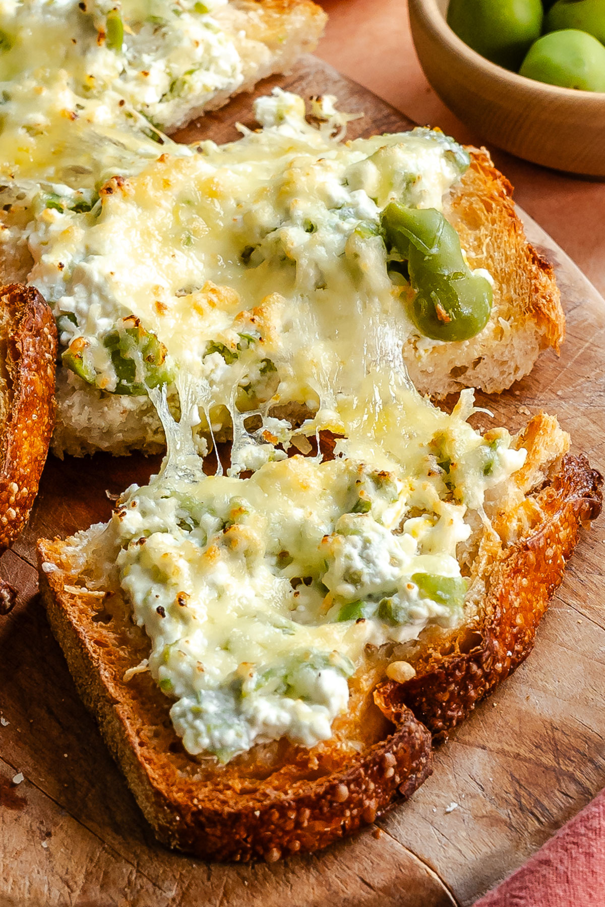 Toasted bread topped with melted cheese, green olives, and ricotta on a wooden board, with a small bowl of green olives in the background.