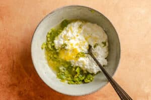 A bowl with cottage cheese, mashed avocado, lemon zest, and a spoon on a light brown surface.