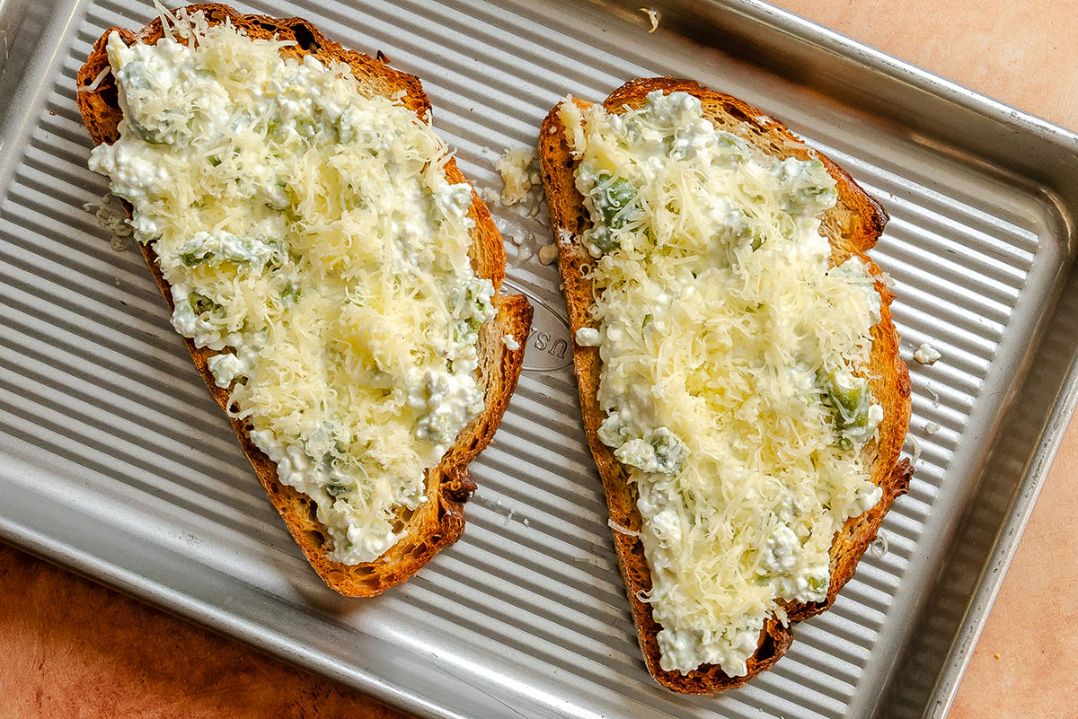 Two slices of bread on a baking tray topped with cottage cheese, herbs, and shredded cheese, ready to be baked.