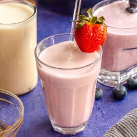 Three glasses of fruit kefir, one with a strawberry garnish, surrounded by blueberries, strawberries, honey, and a gray cloth on a blue surface.
