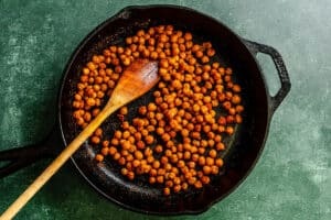 A cast iron skillet contains roasted chickpeas with spices, stirred by a wooden spoon, on a green textured surface.