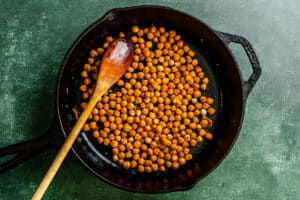 A cast iron skillet with roasted chickpeas and a wooden spoon on a green textured surface.