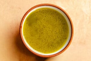A bowl of greenish-yellow soup or broth with an oily surface, placed on a light beige background.