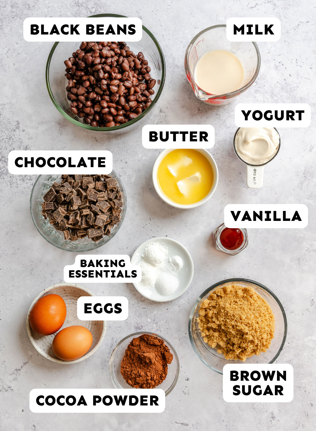 Ingredients for baking no flour black bean muffins, including black beans, milk, yogurt, chocolate, butter, vanilla, eggs, cocoa powder, brown sugar, and baking essentials arranged on a countertop.