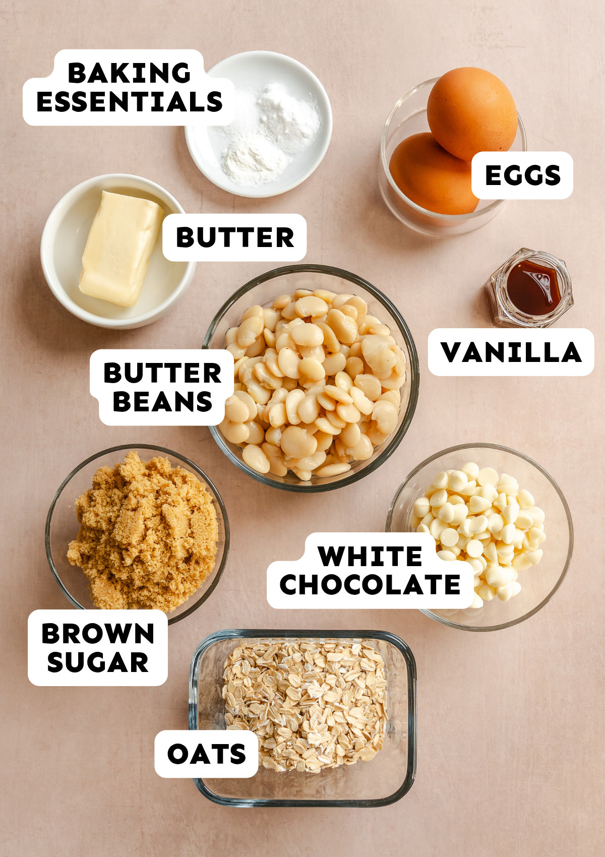 Nine labeled bowls and containers containing the ingredients for Butter Bean Blondies—including baking powder, eggs, butter, vanilla, butter beans, brown sugar, white chocolate chips, and oats—are artfully arranged on a light surface.