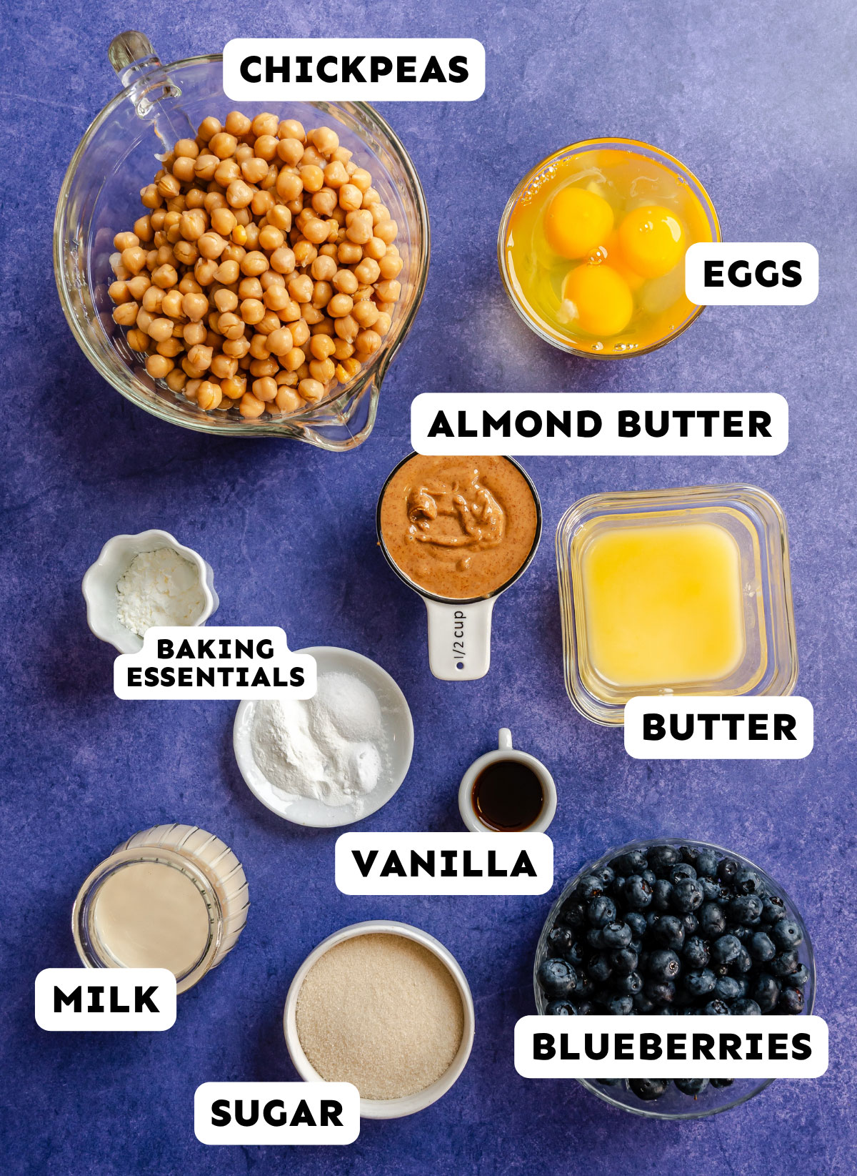 A selection of labeled baking ingredients on a blue surface, perfect for making Chickpea Muffins—including chickpeas, eggs, almond butter, butter, baking essentials, vanilla, milk, sugar, and blueberries.