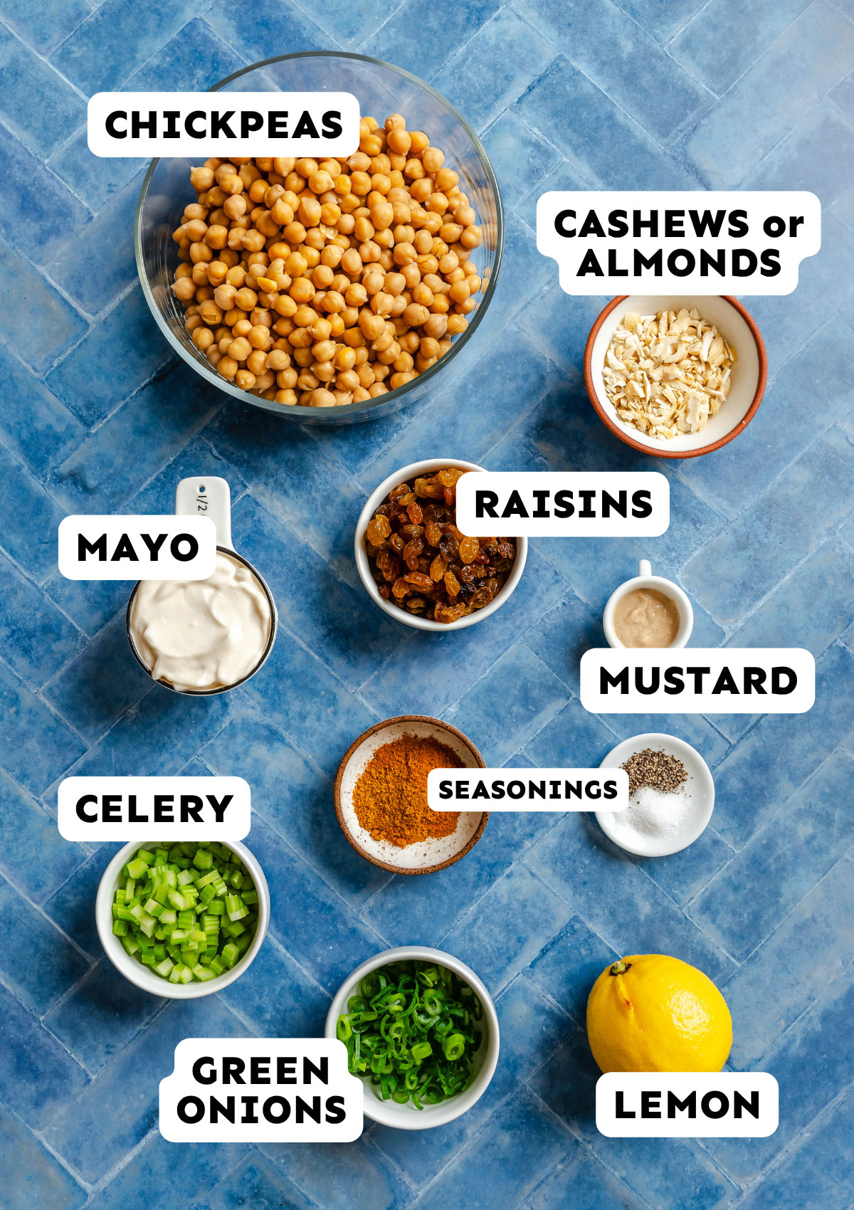 Bowls of chickpeas, cashews or almonds, mayo, raisins, mustard, celery, green onions, seasonings, and a lemon arranged on a blue tiled surface.