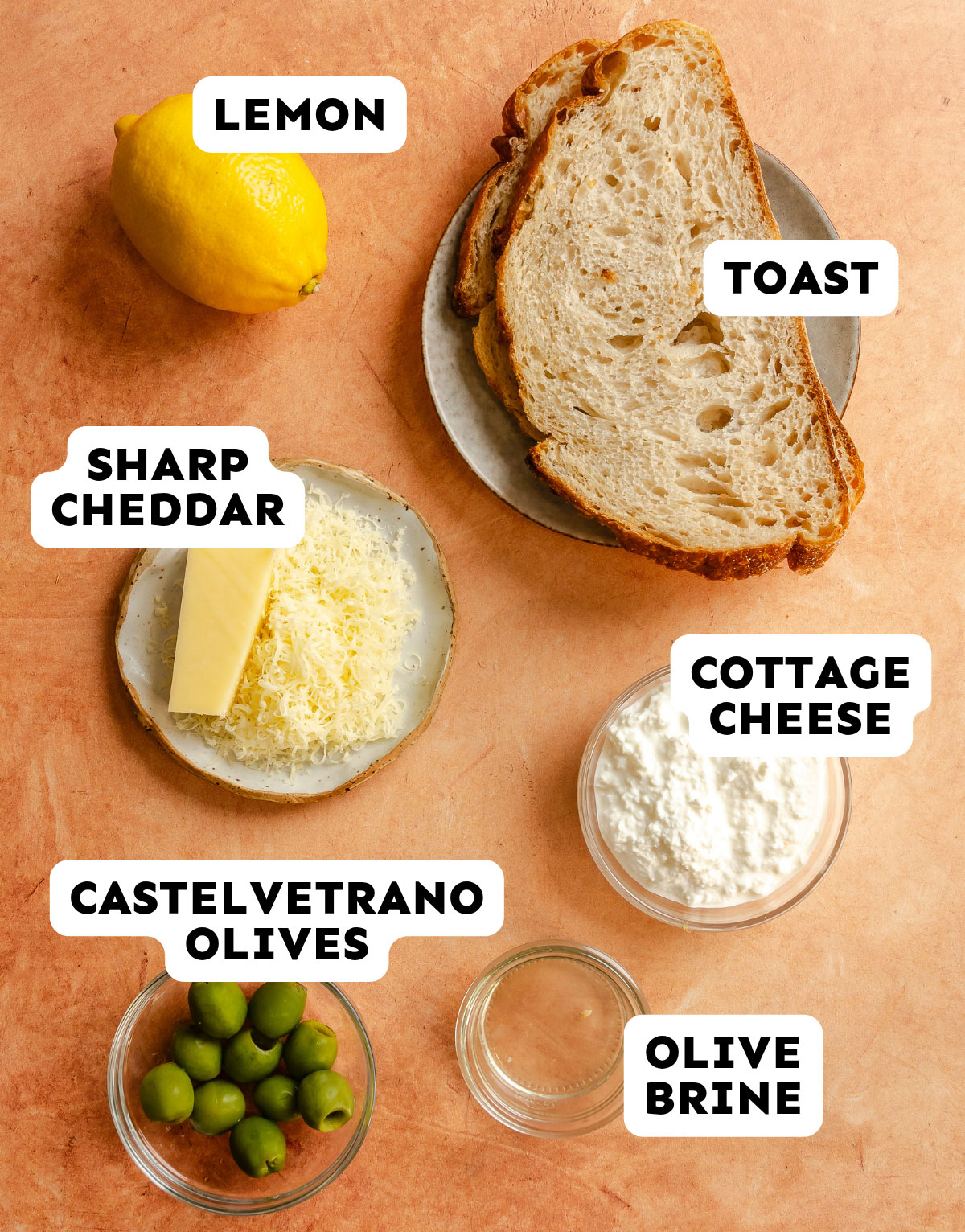 Ingredients on a table: lemon, two slices of toast, grated sharp cheddar, cottage cheese, Castelvetrano olives, and a small bowl of olive brine.