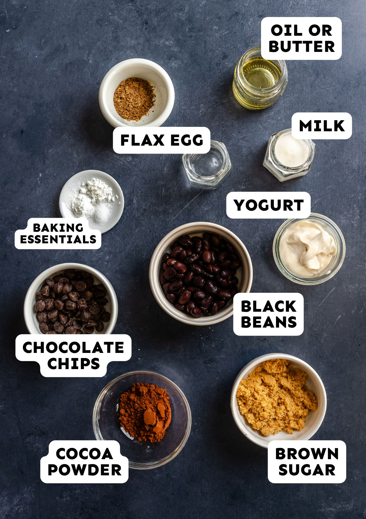 Various labeled bowls contain flax egg, oil or butter, milk, yogurt, baking essentials, black beans—perfect for a decadent Chocolate Mug Cake—chocolate chips, cocoa powder, and brown sugar, all beautifully arranged on a dark surface.