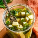 A glass jar filled with sliced pickles, fresh dill, and brine, with a fork resting inside the jar.
